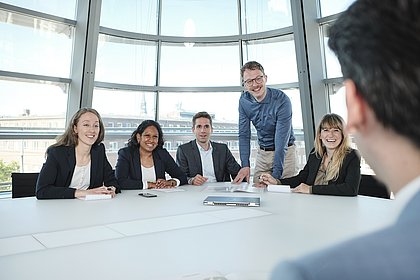 Gruppenaufnahme des Teams vor einem Laptop, im Anschnitt ein weiterer Kollege am Tisch